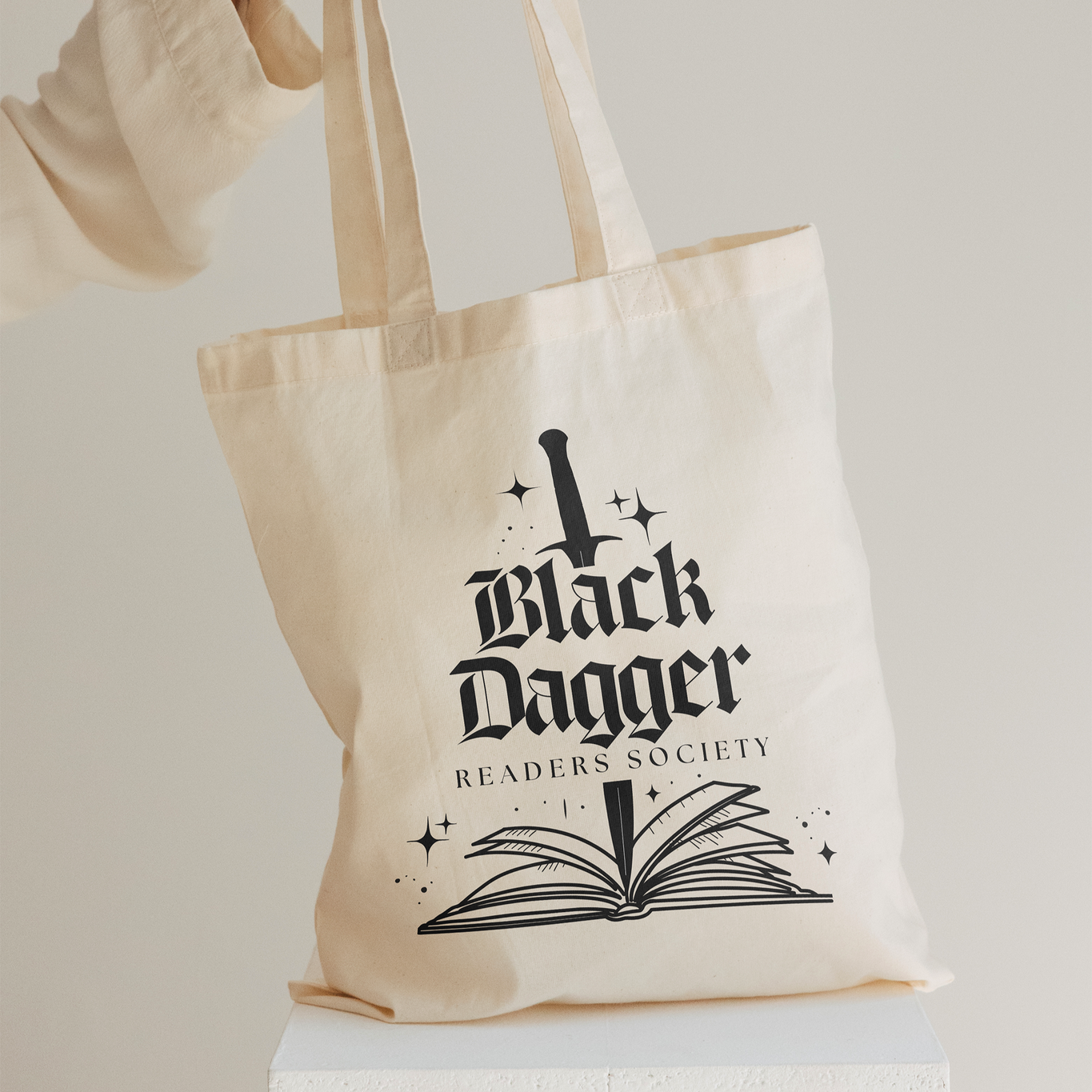 A woman's hand holds a beige tote bag. The tote bag features a black dagger stabbing an open book with the words "Black Dagger Readers Society"