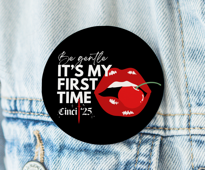 A black round button is pinned to a jean jacket. The button reads "Be gentle it's my first time. Cinci '25" Red lips biting a red cherry are next to the words.