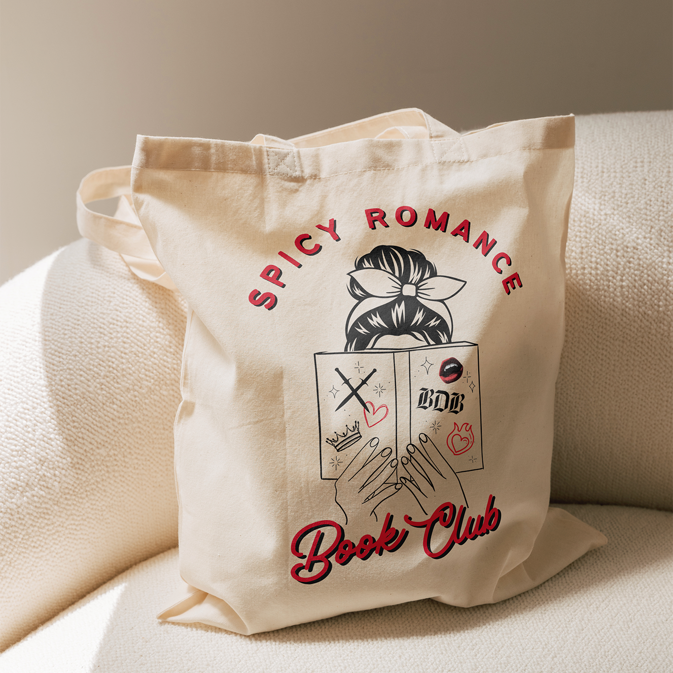 a canvas tote bags sits on a beige chair. The tote bag reads "Spicy Romance Book Club" An image of a woman reading a book is featured between the text.
