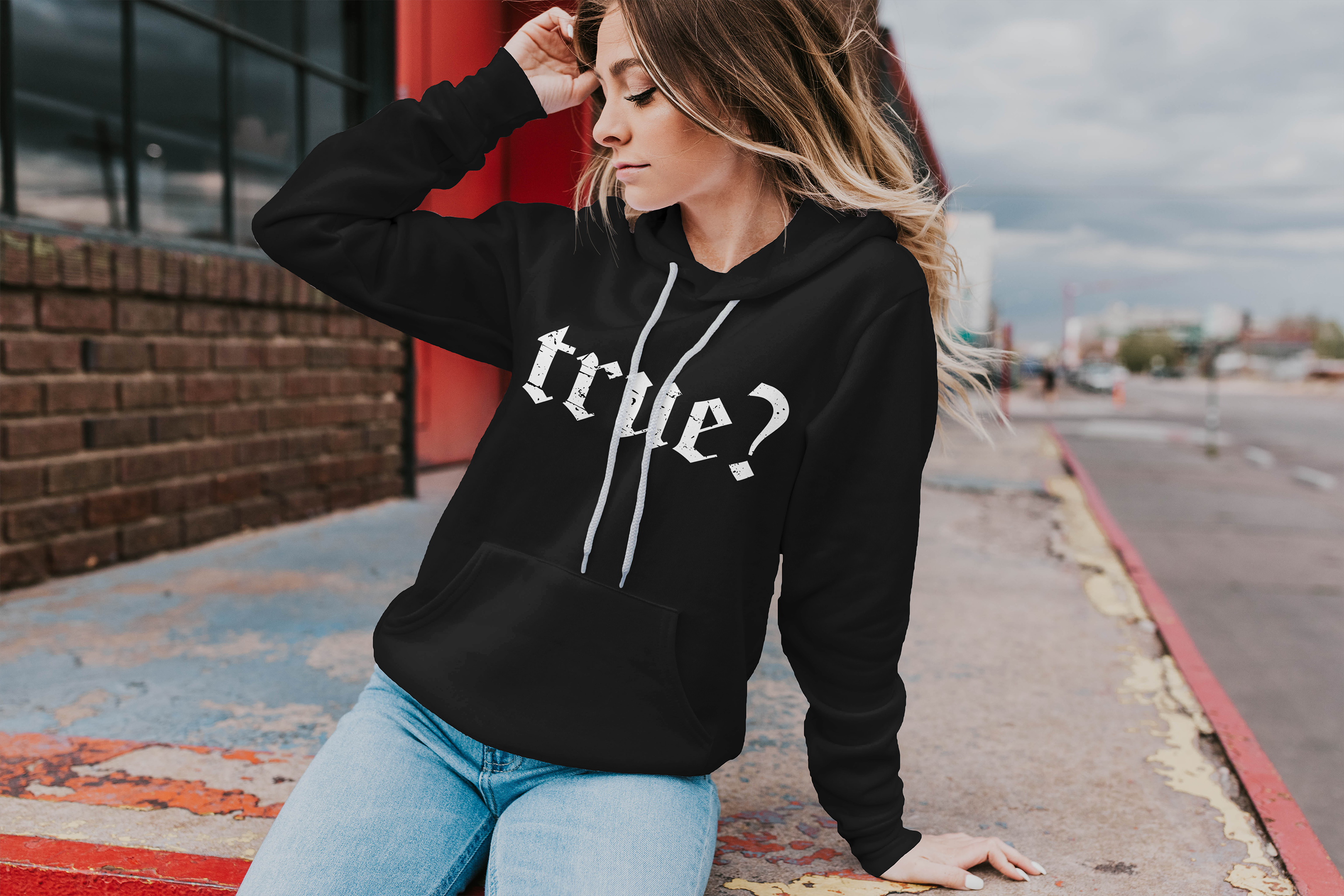 a young woman sits on a concrete edge. She is wearing a black hoodie with the word "True?" on the chest. The hoodie has white strings.
