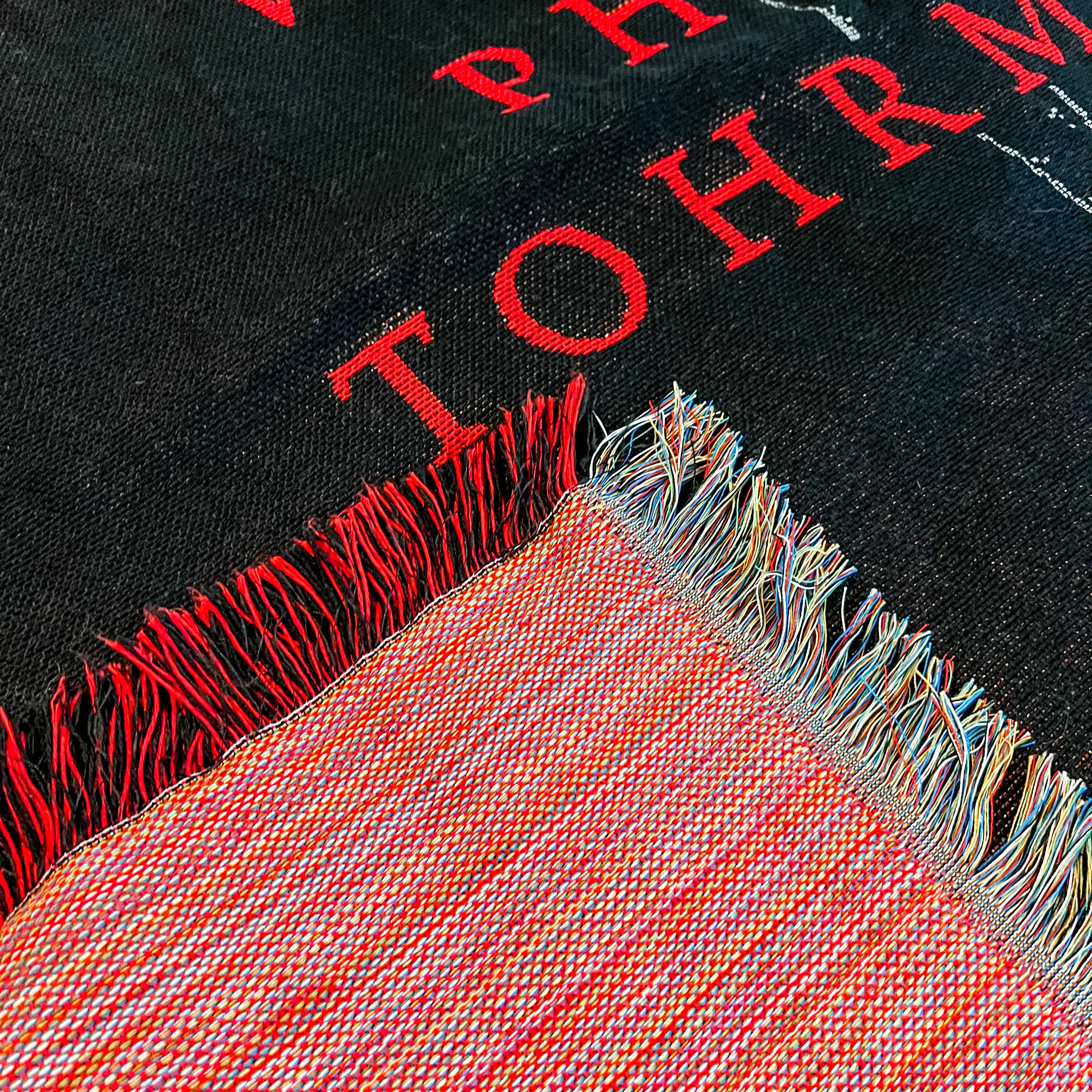Close-up of a black fabric with red text and a fringed mulit-color border.