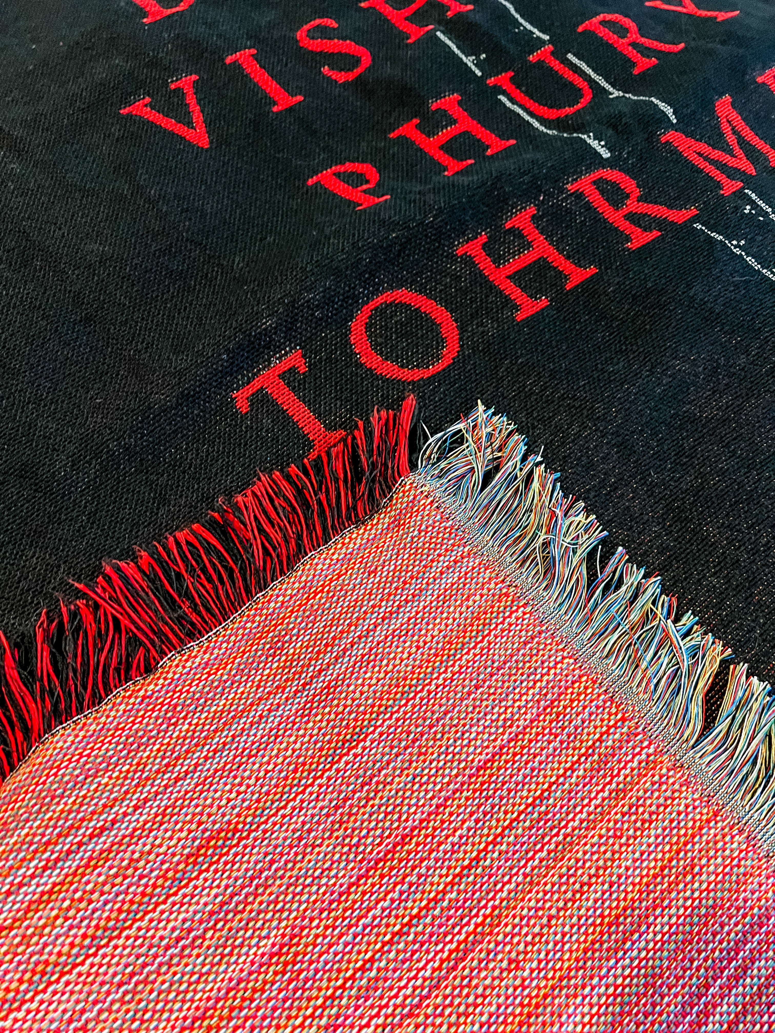 Close-up of a black fabric with red text and a fringed mulit-color border.