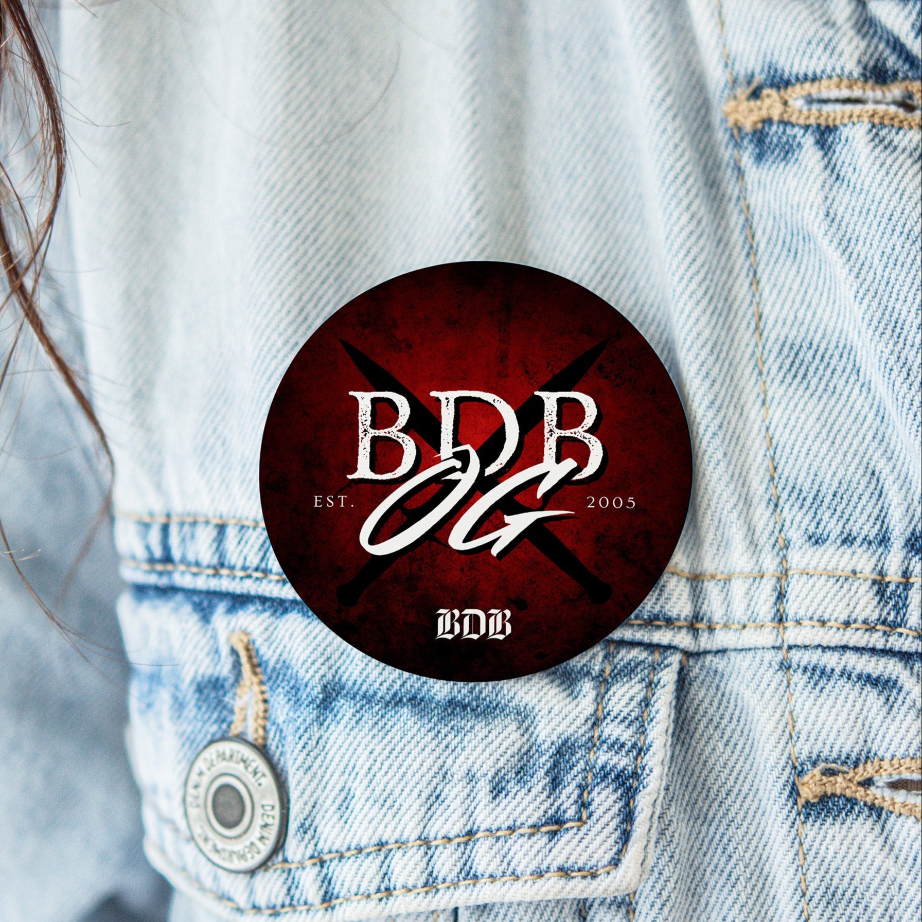 a red button with crossed black daggers with white text overlayed "BDB OG est. 005"