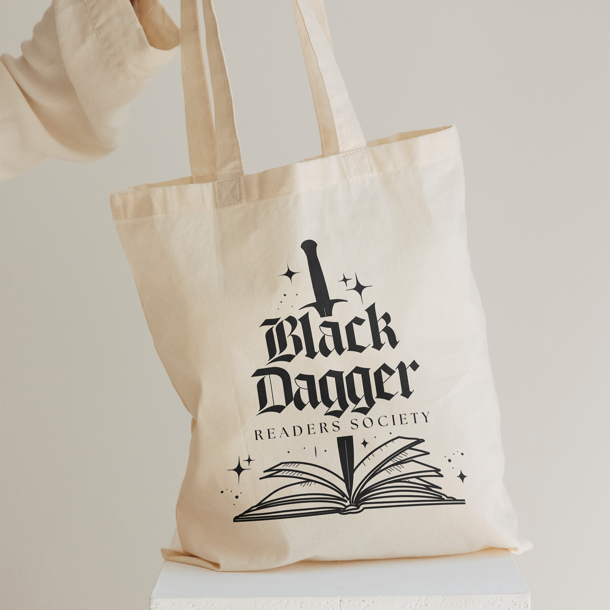 A woman's hand holds a beige tote bag. The tote bag features a black dagger stabbing an open book with the words "Black Dagger Readers Society"