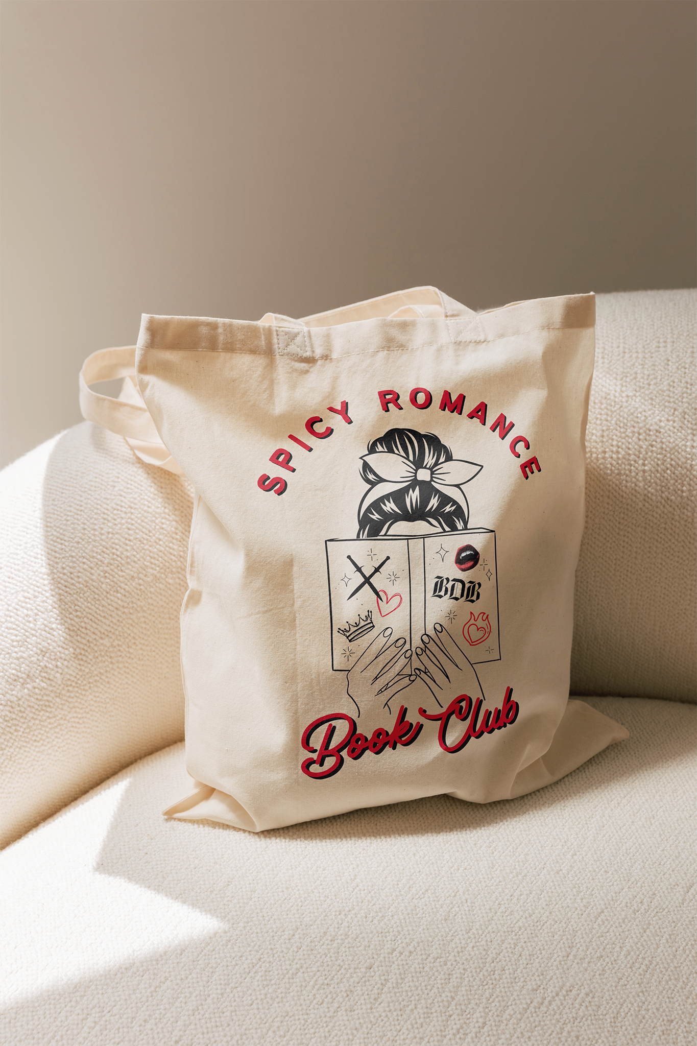 a canvas tote bags sits on a beige chair. The tote bag reads "Spicy Romance Book Club" An image of a woman reading a book is featured between the text.